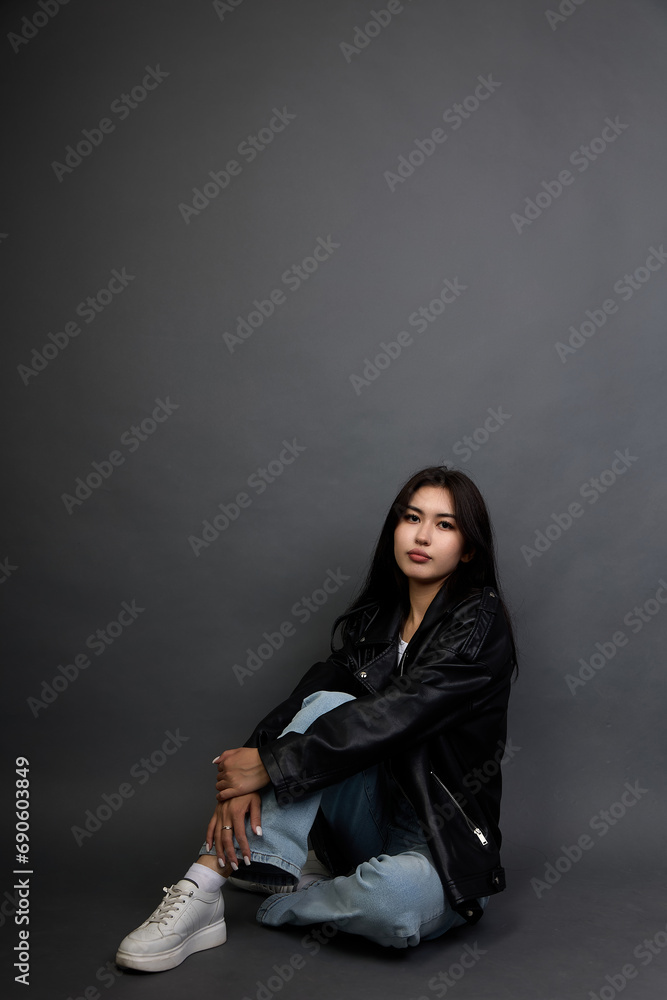 Cute brunette girl with a serious calm expression posing in a black jacket and jeans. Young woman with long dark hair, sitting, hugging her leg with her hand, looking at the camera Cute brunette girl with a serious calm expression posing in a black jacket and jeans. Young woman with long dark hair, sitting, hugging her leg with her hand, looking at the camera