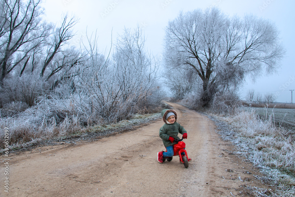 Little child playing outside in frosty winter land on the natural road with toddler motorcycle, frozen woods and twigs nature around