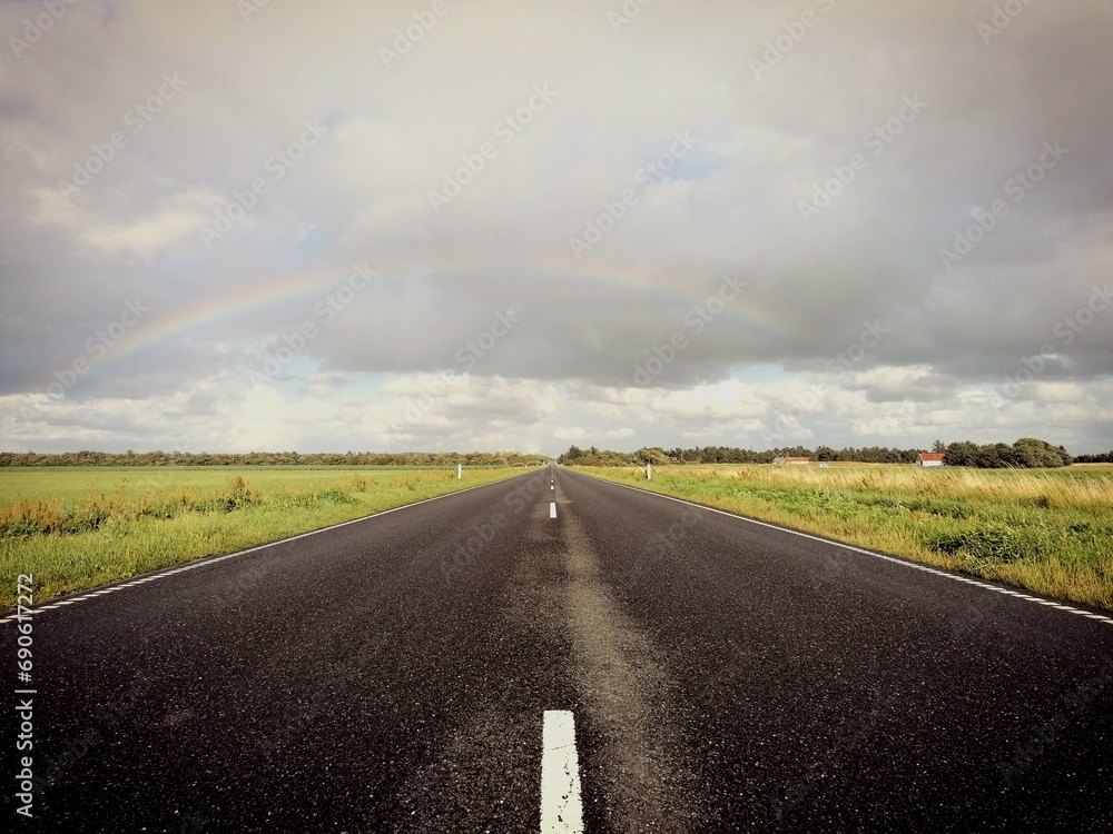 Fototapeta premium Leere gerade Straße mit ländlicher Landschaft zwischen Feldern und Regenbogen