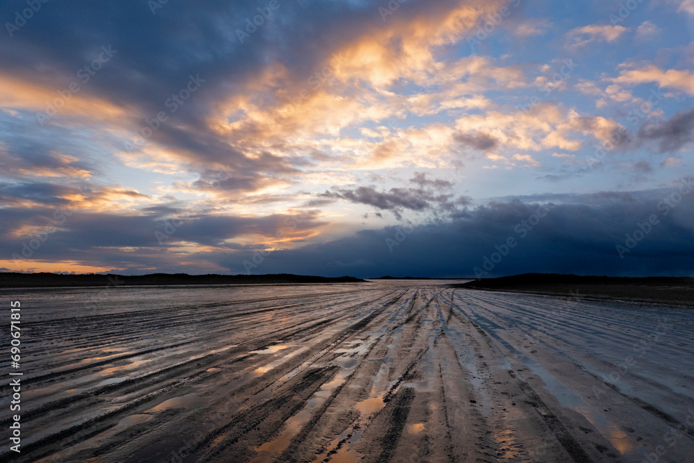 Traces of car tires on a muddy beach, leading to the horizon at nightfall, under a beautifully colored sky, threatening clouds in the distance