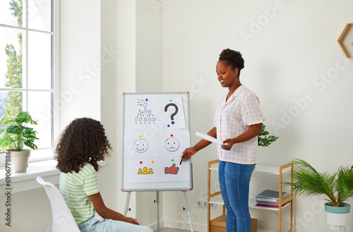 School counselor or psychologist working with a little girl, standing by a white board with pictures, pointing at a thumbs up symbol and asking the child questions. Children's psychology concept photo