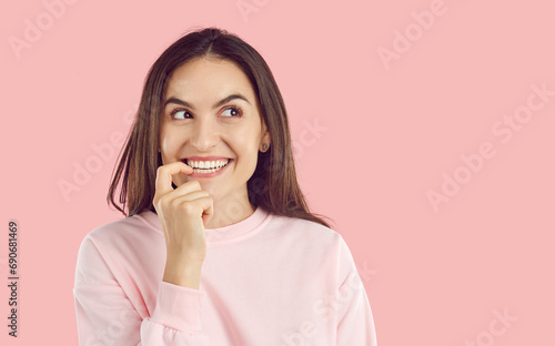 Happy young woman standing on light pink background, looking to copy space side, thinking and smiling. Pretty brunette girl imagines something and looks aside with funny, cheerful, sly face expression