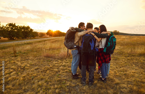 Team of huddling, hugging hikers. Group of happy smiling male and female friends with backpacks standing in circle in autumn field on sunrise or sunset. Tourism, hiking, friendship, teamwork concept