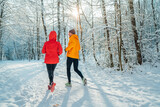 Middle-aged smiling trail couple runners man and woman dressed bright windproof jackets running speaking picturesque snowy forest during sunny frosty day. Sporty active people, winter training image.