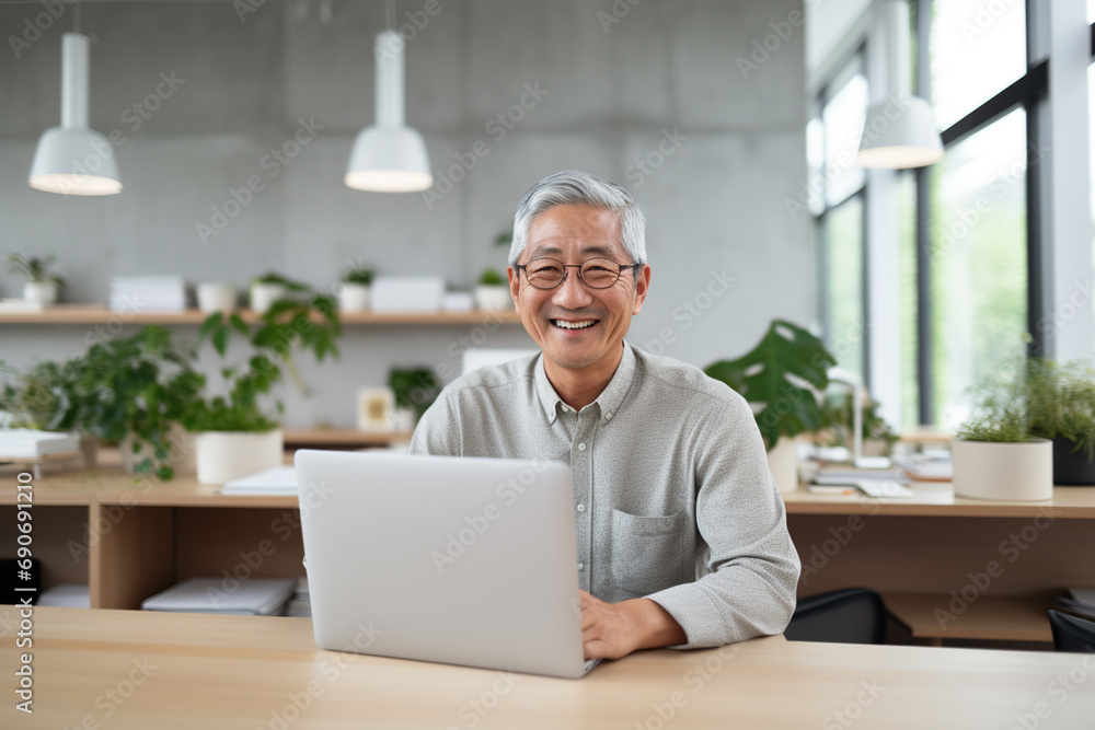Surrounded by a minimalist, bright and calm office space, a 60-year-old Japanese man working on his laptop and grins at the camera Surrounded by a minimalist, bright and calm office space, a 60-year-old Japanese man working on his laptop and grins at the camera