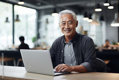 Surrounded by a minimalist, bright and calm office space, a 60-year-old Japanese man working on his laptop and grins at the camera photo