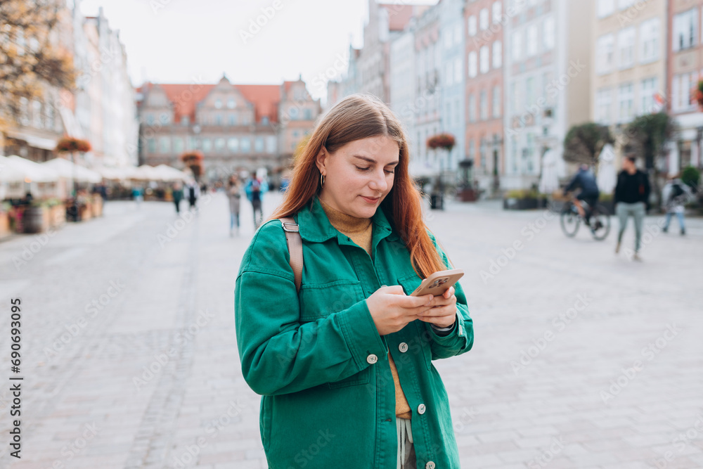 Happy cheerful young woman with backpack walking on city street checks her smartphone. Portrait of beautiful 30s girl using smartphone outdoors. Urban lifestyle concept. Happy cheerful young woman with backpack walking on city street checks her smartphone. Portrait of beautiful 30s girl using smartphone outdoors. Urban lifestyle concept.