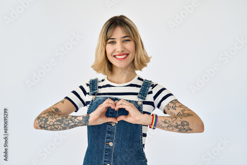 Happy pretty gen z blonde young woman, smiling girl with short blond hair tattoos wearing striped t-shirt denim dress rainbow bracelet showing heart sign standing isolated on white. Portrait. photo