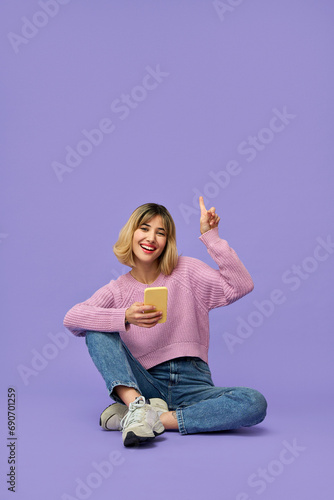 Happy pretty gen z blonde young woman model holding smartphone pointing up at copy space, smiling girl using mobile apps on cell phone sitting isolated on purple background with cellphone, vertical. © insta_photos