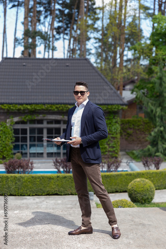 Entrepreneur with sunglasses and clipboard standing in garden of suburban mansion. Realtor prepares for demonstration of house
