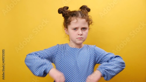 Portrait of little grumpy caucasian girl child pouting lips, grimacing and looking angrily to camera, posing isolated on plain yellow color background wall in studio. People emotion lifestyle concept