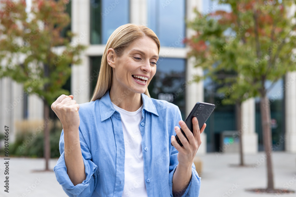Fototapeta premium Happy successful winner woman in blue casual shirt received online notification message on phone, mature business woman outside office building celebrating triumph, joyfully holding hand up.