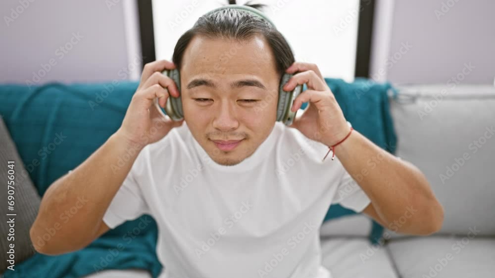 Confident young chinese man radiates joy, removing headphones after enjoying funny song at home, basking in the warmth of his living room atmosphere & smiling energetically Confident young chinese man radiates joy, removing headphones after enjoying funny song at home, basking in the warmth of his living room atmosphere & smiling energetically