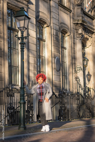Female model with strong unique style appearance in urban wealthy inner city street lamp iron fence environment. Confident curvy black lady stylish red hair with winter good taste fashion