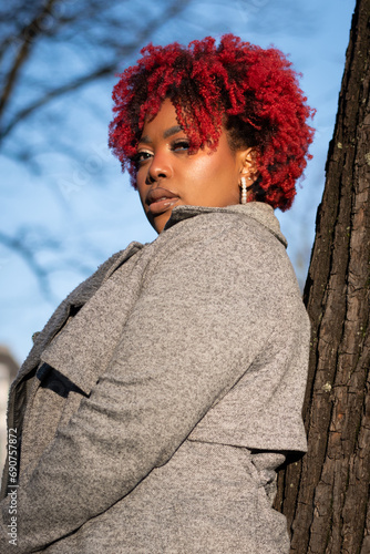 Female model with strong unique style appearance serious moody expression in woodland environment leaning against tree. Confident black lady stylish red hair with winter good taste fashion © drew