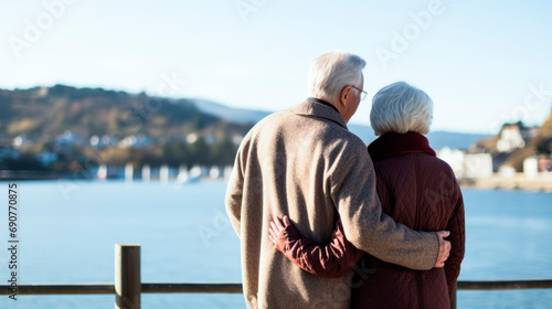 an elderly couple, a man and a senior woman with their arms around each other, look dreamily at the lake, shot from behind photo