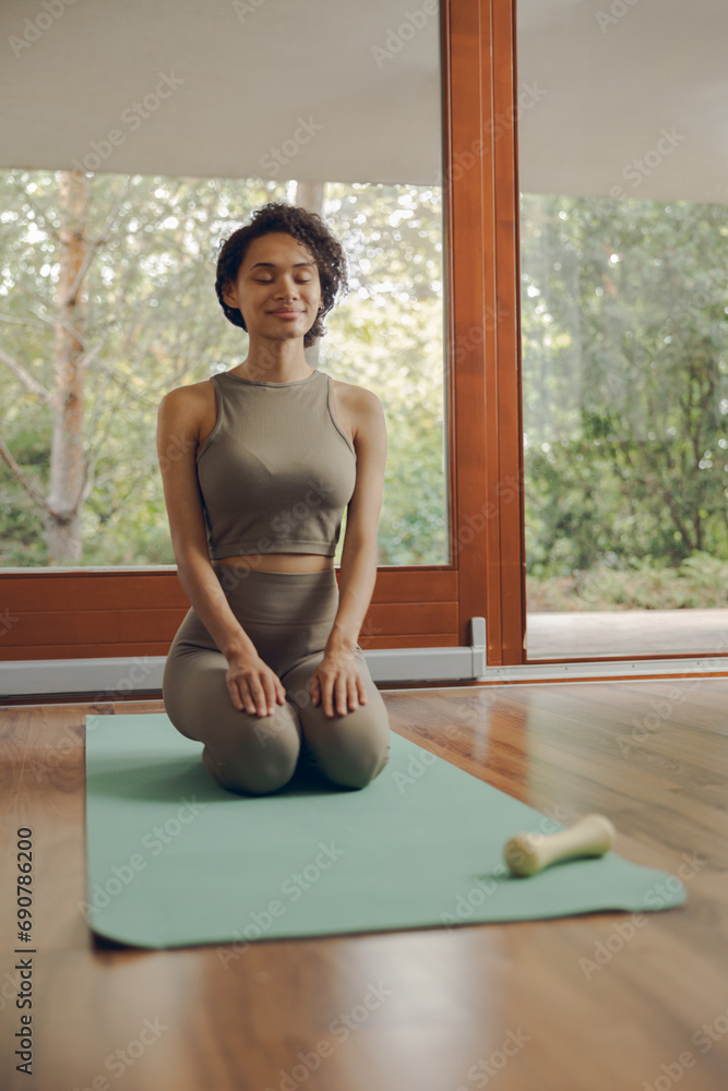 Young smiling woman in activewear sitting on mat at home before doing fitness exercises