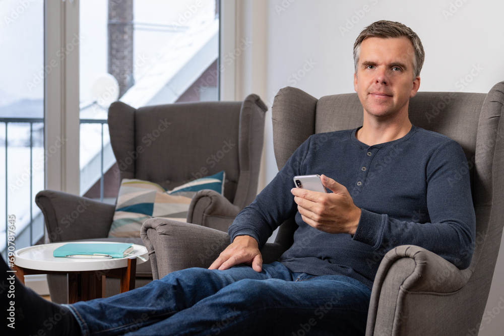 handsome 40s single man sits in an comfy grey armchair with his smartphone in his hand and looks confident into camera
handsome 40s single man sits in an comfy grey armchair with his smartphone in his hand and looks confident into camera