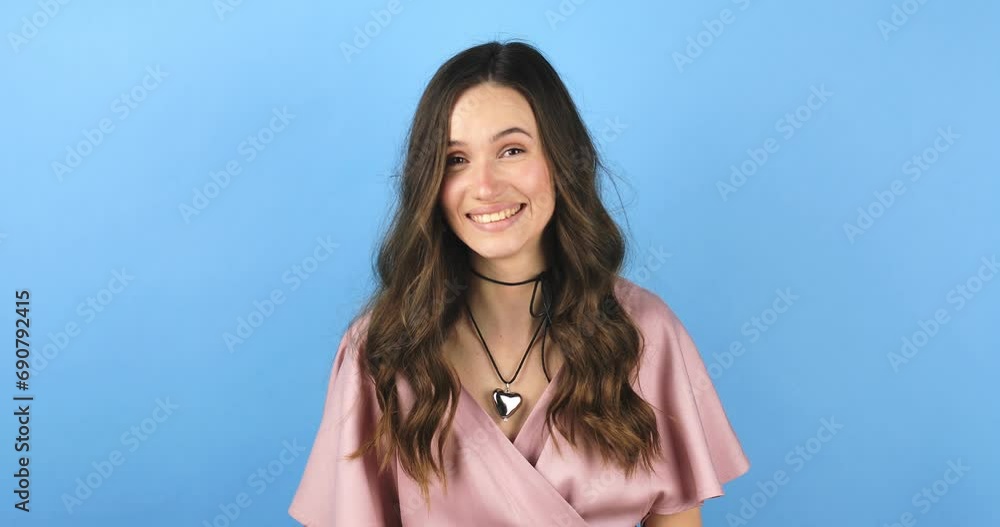 Playful happy contented brunette woman in pink dress blinking eye, looking at camera with toothy smile, winking and flirting, expressing optimism. Indoor studio shot isolated on blue background. Playful happy contented brunette woman in pink dress blinking eye, looking at camera with toothy smile, winking and flirting, expressing optimism. Indoor studio shot isolated on blue background.