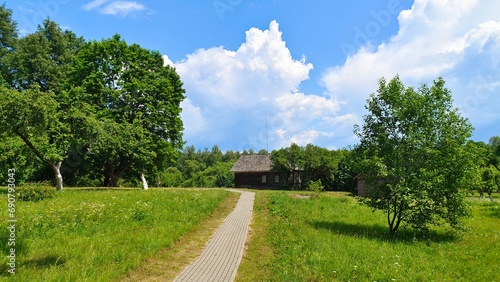 An old wooden house with a tiled roof and lightning rod sits next to an orchard with whitewashed trunks. A tiled path leads to the house through a grassy lawn. Sunny summer weather and blue skies