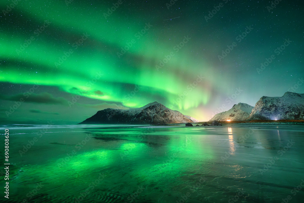 Northern Lights and sandy beach at starry winter night. Lofoten islands, Norway. Beautiful Aurora borealis. Sky with polar lights. Landscape with aurora, sea, sky reflection in water, snowy mountains