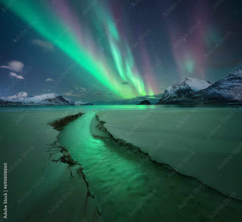 Northern Lights, snowy mountains, sandy beach, creek at starry winter night. Lofoten islands, Norway. Amazing Aurora borealis. Sky with polar lights. Landscape with aurora, sea, stream, rocks in snow