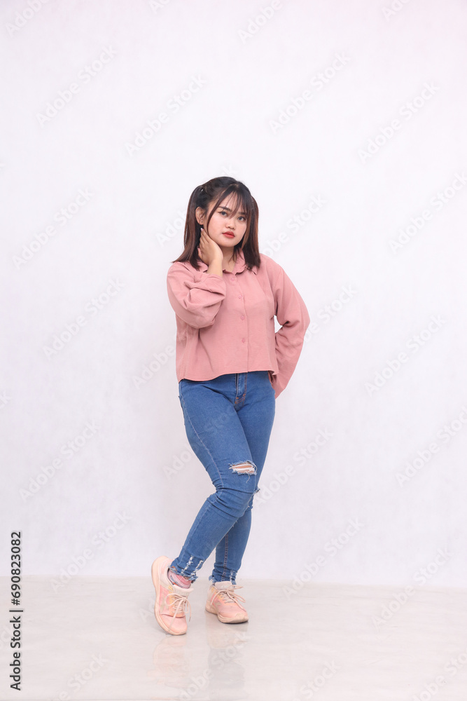 Smiling cheerful black haired beautiful woman in her 20s wearing basic pink shirt standing working style while holding neck and hips white background studio portrait for service promotion Smiling cheerful black haired beautiful woman in her 20s wearing basic pink shirt standing working style while holding neck and hips white background studio portrait for service promotion