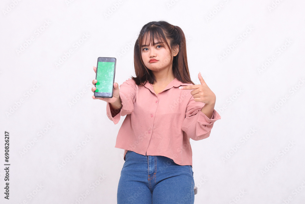 Beautiful happy Asian woman in her 20s wearing casual shirt holding white screen cellphone while frowning pointing at gadget on white background studio portrait for banner ad, banner, billboard Beautiful happy Asian woman in her 20s wearing casual shirt holding white screen cellphone while frowning pointing at gadget on white background studio portrait for banner ad, banner, billboard