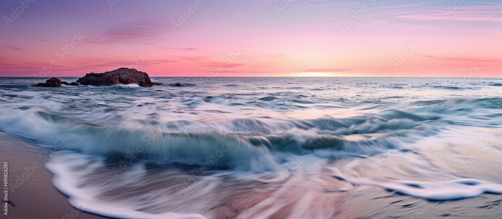Obraz premium The colorful sunset is seen from the blue, pink sea beach with the waves gently flowing onto the shore