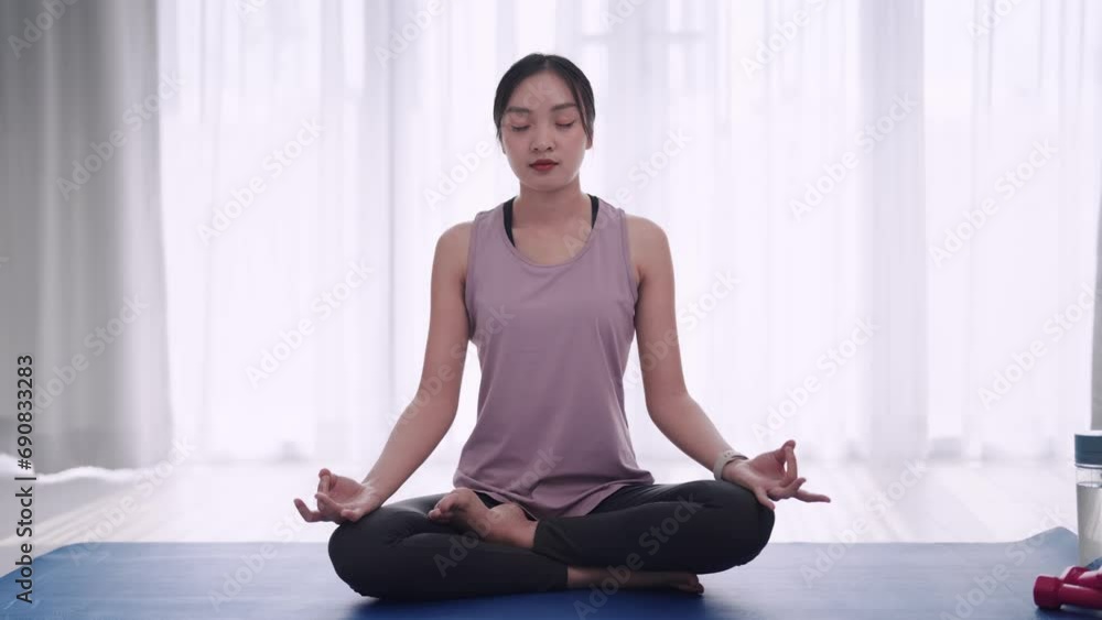 Asian woman in workout attire practices meditation on a yoga mat in her home's sitting room. Perfect for showcasing relaxation and mindfulness in a domestic setting. Asian woman in workout attire practices meditation on a yoga mat in her home's sitting room. Perfect for showcasing relaxation and mindfulness in a domestic setting.
