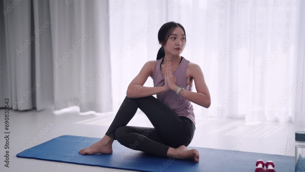 An Asian woman in workout attire gracefully stretches her muscles using yoga or exercise poses on a mat in her home's sitting room. Ideal for fitness or yoga promotion An Asian woman in workout attire gracefully stretches her muscles using yoga or exercise poses on a mat in her home's sitting room. Ideal for fitness or yoga promotion