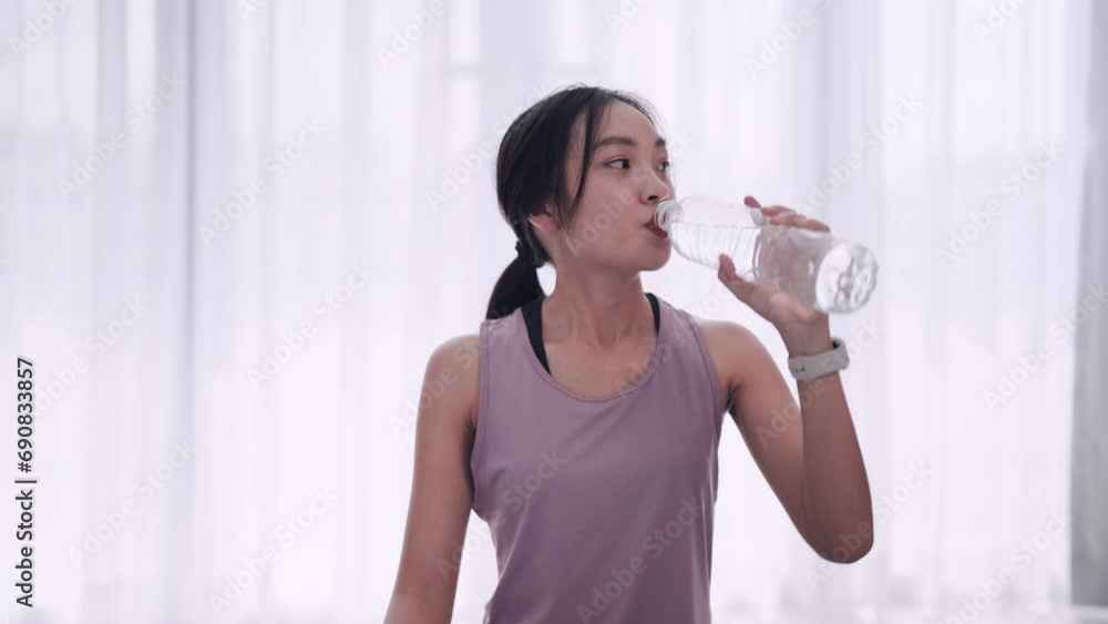 An Asian woman in workout attire takes a refreshing drink after exercising on a mat in her home's sitting room. Ideal for promoting health and fitness, showcasing post-workout hydration An Asian woman in workout attire takes a refreshing drink after exercising on a mat in her home's sitting room. Ideal for promoting health and fitness, showcasing post-workout hydration