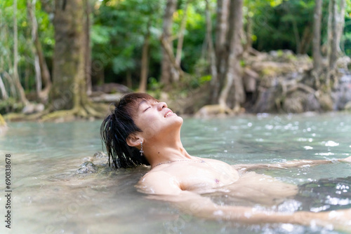 Young Asian man travel nature on summer holiday vacation. Happy generation z guy relax and enjoy outdoor lifestyle jumping and playing water at waterfall lagoon in forest mountain.