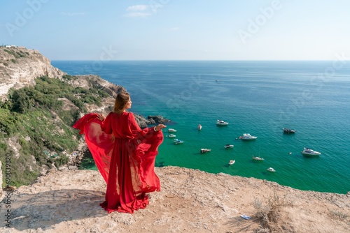 Red Dress Woman sea Cliff. A beautiful woman in a red dress and white swimsuit poses on a cliff overlooking the sea on a sunny day. Boats and yachts dot the background.