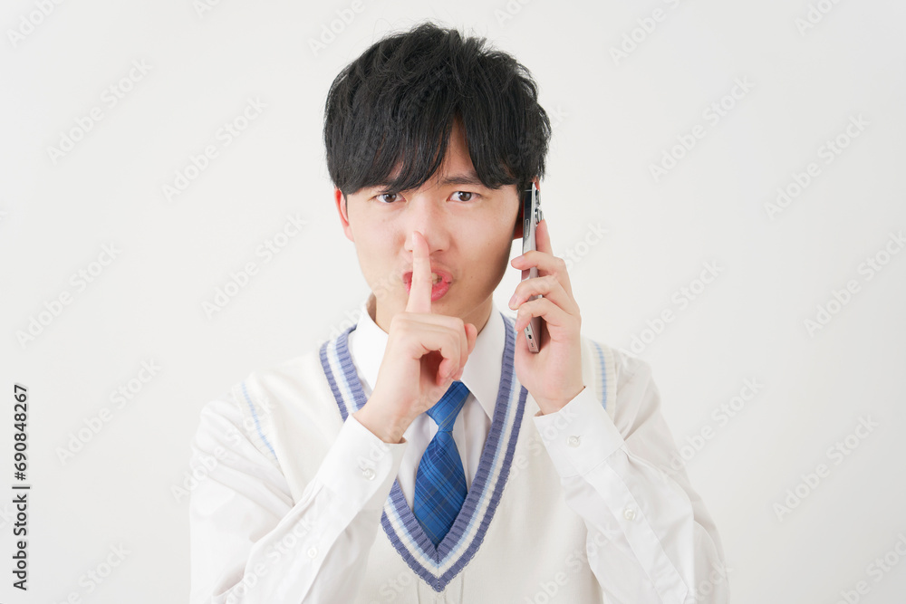 スマートフォンで電話をしながら内緒のポーズをする男子高校生 白背景 スマートフォンで電話をしながら内緒のポーズをする男子高校生 白背景