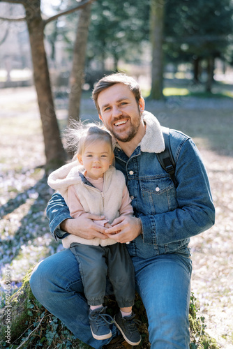 Happy dad with a little girl on his knees sit on a stump in a spring park © Nadtochiy