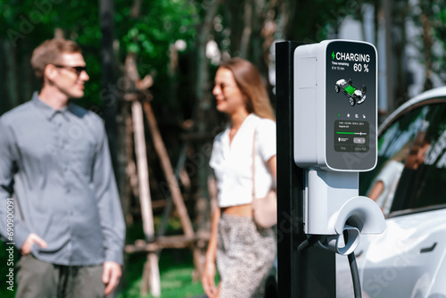 Young couple travel with EV electric car charging in green sustainable city outdoor garden in summer shows urban sustainability lifestyle by green clean rechargeable energy of electric vehicle innards