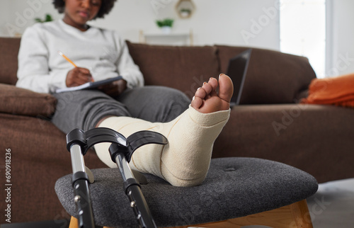 African American woman with broken leg sitting on sofa at home and writing in diary, with her injured foot resting on stool, with black crutches beside. Foot wrapped in bandage closeup. Injury concept photo