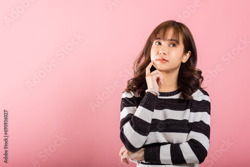 In a studio shot on a pink background, an Asian woman stands with her chin in her hand, showing a thoughtful expression as she ponders a question. photo