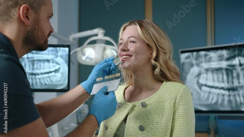 Cute blonde woman standing smiling while young dental expert holding samples of implants choosing suitable shade for veneers. Clinic with modern equipment. High quality 4k footage photo