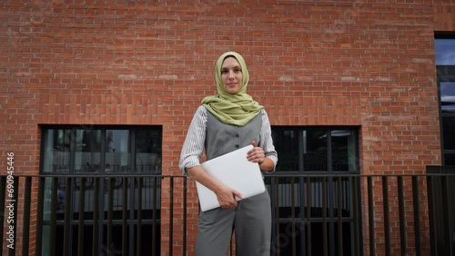 Beautiful woman in hijab standing on city street. Muslim businesswoman with laptop in hands. Iran, Afganistan female teacher in front of school building. photo