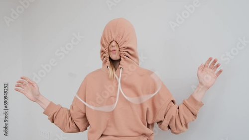 Portrait of a young teenage woman with a large birthmark on her face in a peach beige hoodie tightens the hood with a drawstring, hiding and then revealing knot looking at camera on a white background photo