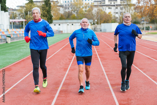 Three elderly Caucasian men in blue gear jog together on a cloudy day at an outdoor track