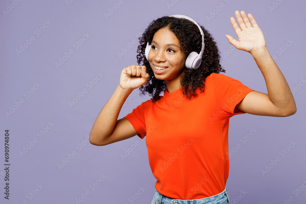 Fototapeta premium Little kid teen girl of African American ethnicity wear orange t-shirt listen to music in headphones dance raise up hands isolated on plain pastel purple background studio Childhood lifestyle concept