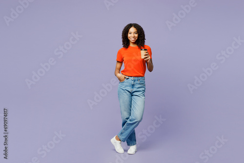 Full body little kid teen girl of African American ethnicity wear orange t-shirt hold takeaway delivery craft paper cup coffee to go isolated on plain purple background. Childhood lifestyle concept.