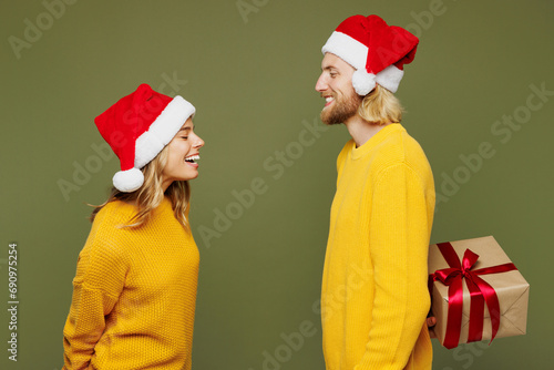 Side view merry young couple two friend man woman wear sweater Santa hat posing give present box with gift ribbon bow isolated on plain green background. Happy New Year celebration Christmas concept.