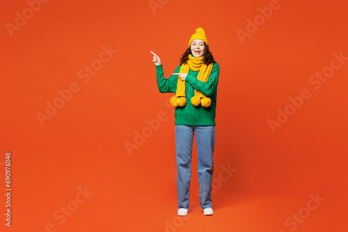 Full body young smiling cheerful happy woman she wear green knitted sweater yellow hat scarf point index finger aside on area isolated on plain orange red background studio portrait Lifestyle concept