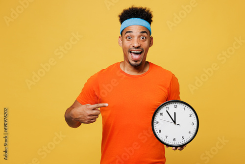 Young amazed fitness trainer instructor sporty man sportsman wear orange t-shirt hold in hand point on clock spend time in home gym isolated on plain yellow background. Workout sport fit abs concept. photo