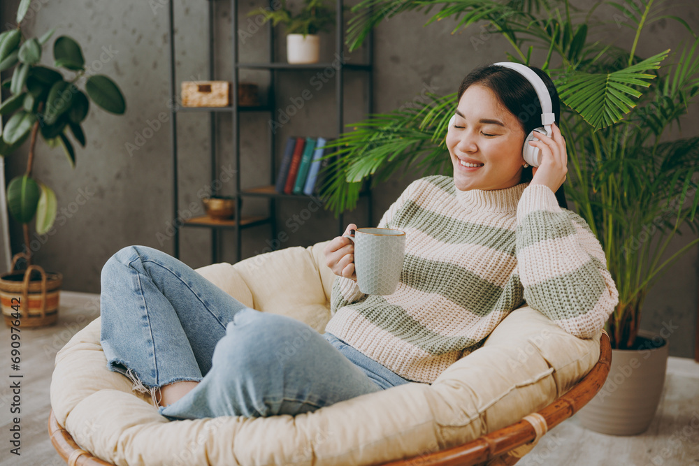Obraz premium Side view young woman wears casual clothes listen to music in headphones drink tea sits on armchair stay at home hotel flat rest relax spend free spare time in grey living room indoor. Lounge concept.