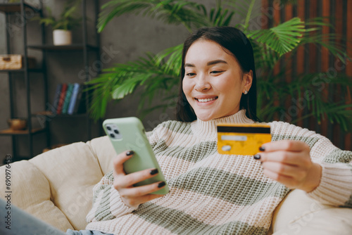 Young woman in casual clothes sits on armchair hold credit card use mobile cell phone shopping online stay home hotel flat rest relax spend free spare time in grey living room indoor. Lounge concept. photo